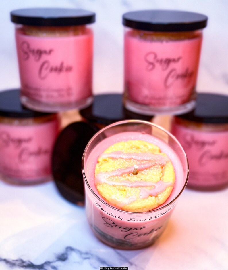 Peut inclure: Cinq pots de bougies en verre rose avec des couvercles noirs. Les pots de bougies sont &eacute;tiquet&eacute;s "Sugar Cookie" et ont un biscuit au sucre rose sur le dessus de la bougie dans le pot du centre.