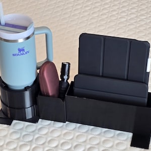 May include: A black car organizer with a light blue Stanley tumbler, a maroon item, a black flashlight, and a black tablet case. The tumbler has a handle and a straw. The organizer has a cup holder and storage compartments.
