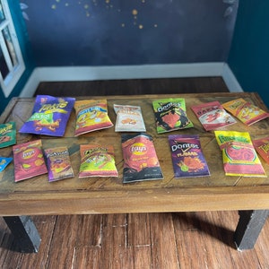 Może przedstawiać: Kolekcja miniaturowych torebek ze przekąskami na drewnianym stole. Torebki przedstawiają różne popularne marki, takie jak Takis, Lays, Doritos i Funyuns. Torebki są w różnych kolorach i mają różne wzory.