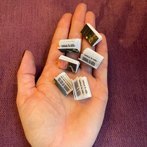 K&ouml;nnte beinhalten: Eine Hand h&auml;lt eine Sammlung von Miniaturb&uuml;chern. Die B&uuml;cher sind geschlossen und haben verschiedene Titel und Umschl&auml;ge.