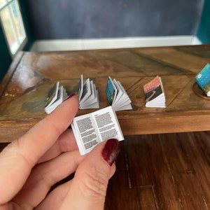 K&ouml;nnte beinhalten: Eine Sammlung von Miniaturb&uuml;chern auf einem Holztisch. Die B&uuml;cher sind ge&ouml;ffnet und zeigen verschiedene Illustrationen und Texte. Ein Buch wird in einer Hand gehalten.