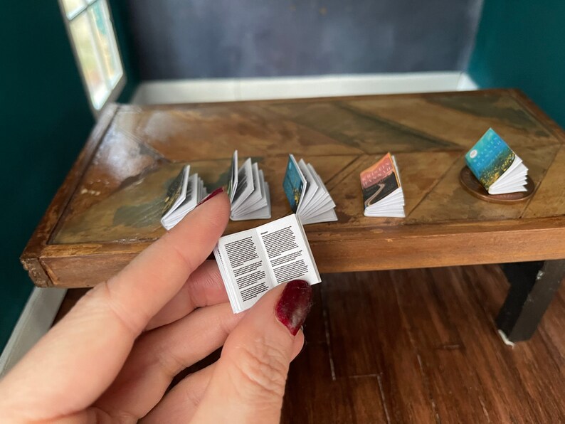 K&ouml;nnte beinhalten: Eine Sammlung von Miniaturb&uuml;chern auf einem Holztisch. Die B&uuml;cher sind ge&ouml;ffnet und zeigen verschiedene Illustrationen und Texte. Ein Buch wird in einer Hand gehalten.