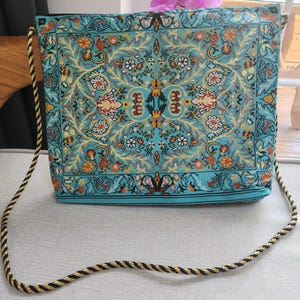 May include: A turquoise shoulder bag with a detailed floral pattern in shades of yellow, orange, and brown. The bag has a black trim and a rope strap with black and gold threads. The bag is rectangular in shape.