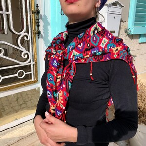 May include: A black mock neck shirt with a colorful patterned scarf. The scarf has a geometric design with red, blue, green, and yellow accents. The scarf is tied around the neck and hangs down the front of the shirt.
