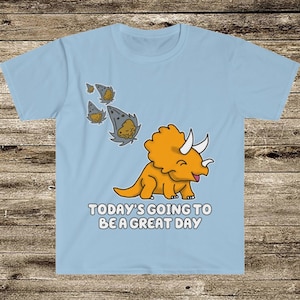 May include: A light blue t-shirt with a cartoon illustration of a happy orange triceratops dinosaur. The dinosaur is facing right and has three horns on its head. The text "TODAY'S GOING TO BE A GREAT DAY" is printed below the dinosaur. There are five small gray asteroids flying behind the dinosaur.