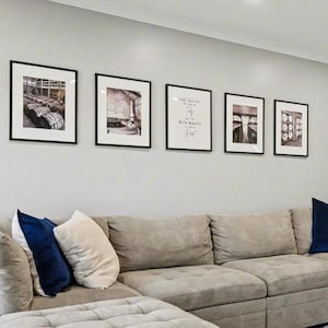 Puede incluir: Un salón moderno con un gran sofá seccional gris claro y una otomana. Cojines decorativos en azul marino y crema añaden contraste. Encima del sofá, cinco grabados en blanco y negro enmarcados cuelgan de la pared gris claro. Una alfombra estampada y pisos de madera completan el espacio.