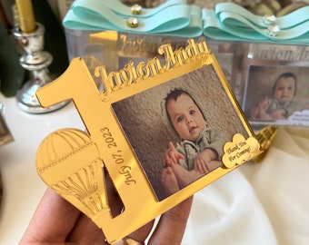 May include: Gold-colored acrylic photo frame with a hot air balloon design. The frame has a picture of a baby boy and the text "Javion Andy" and "July 07, 2023". A small gold-colored tag with the text "Thank You For Coming!" is attached to the frame.