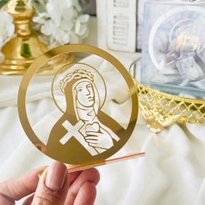 Könnte beinhalten: Ein goldfarbenes, kreisförmiges religiöses Symbol mit einer Darstellung eines Heiligen mit Heiligenschein, Kreuz und Herz. Das Symbol wird vor einem weißen Hintergrund gehalten, mit anderen dekorativen Gegenständen.