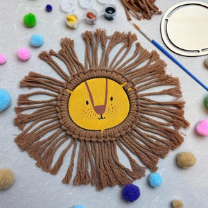 Könnte beinhalten: Ein DIY-Bastelset mit einem Löwen-Design. Das Set enthält ein Holz-Löwengesicht mit gelbem Hintergrund und braunen Details, umgeben von braunem Makramee. Farbtöpfe, ein Pinsel und bunte Pompons sind ebenfalls enthalten.