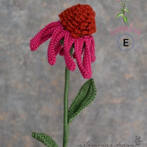 May include: A handmade crochet flower with a vibrant red-orange center and bright pink petals. The stem is green, with a green leaf. The flower is set against a gray background. The text "kineZYA" and "metagurumi_design" are visible.