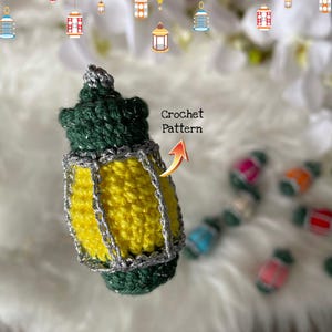 May include: A crocheted green and yellow lantern with silver trim. The lantern is hanging from a string and there are other lanterns in the background. The text "Crochet Pattern" is written on the image.