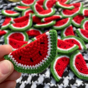 May include: A collection of crocheted watermelon slices in red, green and white. The watermelon slices are arranged in a pile on a black and white checked fabric.