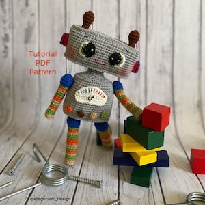 Pode incluir: Um brinquedo de robô de crochê cinza com pernas e braços listrados coloridos. O robô está segurando uma pilha de blocos de madeira coloridos. O texto "Tutorial PDF Pattern" é visível na imagem.