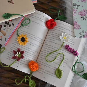 Può includere: Due segnalibri all'uncinetto con motivi floreali. Uno presenta un girasole giallo, un fiore rosso scuro e un fiore arancione. L'altro ha una rosa rossa, un fiore bianco e un piccolo grappolo viola, tutti con steli e foglie di colore verde.