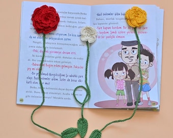 May include: Three crochet flower bookmarks resting on an open book. The bookmarks feature a red rose, a small white flower, and a yellow flower, each attached to a long green stem with crocheted leaves. The book pages contain printed text and illustrations.