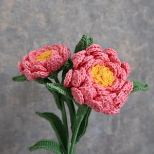 Puede incluir: Un par de flores de ganchillo hechas a mano con pétalos rosas y centros amarillos. Las flores tienen tallos y hojas verdes, sobre un fondo gris texturizado. La artesanía muestra un diseño detallado y texturizado.