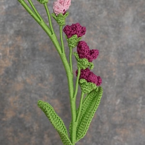 May include: A handmade crochet flower stem with multiple blooms. The flowers are in shades of pink and magenta, with green accents on the buds and leaves. The stem is a vibrant green, and the overall effect is delicate and charming.