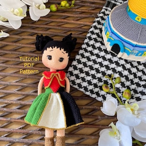 Könnte beinhalten: Gehäkelte Puppe, die ein rot-weiß-grün-schwarzes Kleid mit goldenen Verzierungen trägt. Die Puppe hat schwarze Haare und braune Augen. Die Puppe sitzt in einem geflochtenen Korb mit einem weiß-schwarz karierten Stoff und einer gehäkelten Kuppelstruktur.