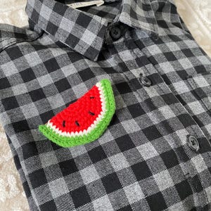 May include: A crocheted watermelon slice pin in red, green, and white. The pin is on a black and grey plaid shirt.