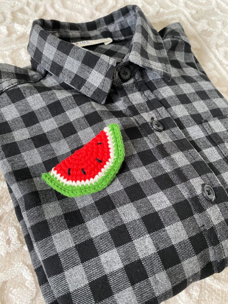 May include: A crocheted watermelon slice pin in red, green, and white. The pin is on a black and grey plaid shirt.