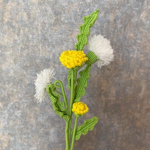 Puede incluir: Ramo de flores de diente de león de ganchillo con cabezas de flores amarillas y blancas, tallos y hojas verdes. Las flores hechas a mano se exhiben sobre un fondo gris texturizado. Un artículo artesanal único.