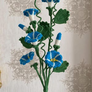 May include: A handcrafted crochet floral arrangement featuring blue and white morning glory flowers with yellow centers, green leaves, and stems. The flowers are detailed with white stripes and are set against a neutral background.