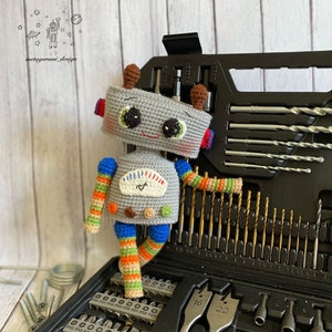 Puede incluir: Un juguete robot de crochet gris con piernas rayadas azules, naranjas y verdes. El robot está de pie en una caja de herramientas negra llena de brocas y puntas de destornillador. La caja de herramientas tiene etiquetas para los diferentes tamaños de brocas, incluyendo "PH0 PH1 PH2 PH3 P20 P21 PZ2 PZ3" y "16 22".