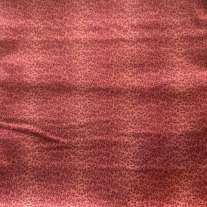May include: Close-up of a fabric with a repeating pattern of small, dark purple leaves on a light red background. The fabric has a slightly wrinkled texture, with visible folds and shadows. The overall color palette is warm and inviting.