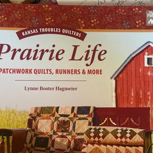 May include: A softcover book titled Prairie Life: Patchwork Quilts, Runners & More by Lynne Boster Hagmeier. The cover features a red barn, a field, and a quilt draped over a wooden bench, with floral patterns at the top and white text.