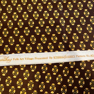 May include: A brown fabric with a repeating pattern of yellow flowers and green leaves. The fabric has a white label that reads "Williamsburg Folk Art Village Presented By WINDHAM FABRICS Pattern No. #31692".