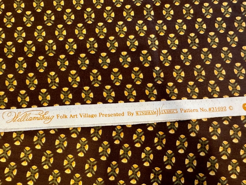 May include: A brown fabric with a repeating pattern of yellow flowers and green leaves. The fabric has a white label that reads "Williamsburg Folk Art Village Presented By WINDHAM FABRICS Pattern No. #31692".