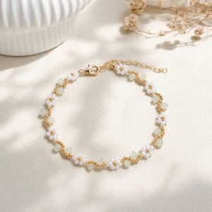 Pode incluir: Uma pulseira delicada com um padrão repetitivo de pequenas contas brancas em forma de flor, contas redondas verde-pálido e minúsculas contas metálicas douradas. A peça inclui um fecho lagosta dourado e uma corrente de elos ajustável.