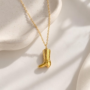May include: A gold-toned necklace featuring a delicate chain and a pendant shaped like a cowboy boot. The boot pendant has a polished finish with intricate embossed detailing along the shaft and a small heel, suspended from a thin link chain.