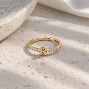 Op de afbeelding: Een goudkleurige verstelbare ring met een centrale letter R en een band bezet met kleine, heldere, fonkelende steentjes. De metalen band heeft een zichtbare opening aan de achterkant, waardoor de maat kan worden aangepast aan verschillende vingers.