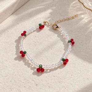 May include: A beaded bracelet featuring clear, faceted glass beads interspersed with small red and green cherry-shaped charms. The bracelet includes a gold-toned metal chain extender and a lobster claw clasp, creating a delicate and colorful accessory.