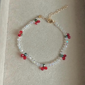 Op de afbeelding: Een delicate armband met heldere kristallen kralen en kleine clusters van rode en groene kralen. De armband heeft een goudkleurige ketting en sluiting. Het ontwerp is eenvoudig en elegant, geschikt voor dagelijks gebruik of speciale gelegenheden.