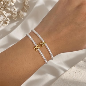 May include: Two delicate bracelets made of small, round, white faux pearls. Each bracelet features a single, polished gold-tone metal letter charm, one displaying the letter K and the other displaying the letter A, strung on a flexible cord.