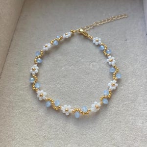 Puede incluir: Un delicado brazalete de cadena dorada con flores de cuentas blancas y azul claro.