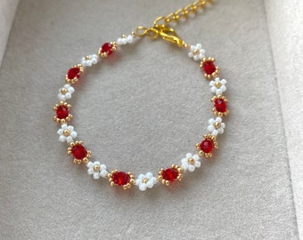 May include: A delicate bracelet made with white and red beads, featuring a gold chain clasp. The bracelet is made of small, flower-shaped beads, with red beads in the center of each flower.