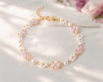 May include: A delicate bracelet featuring a repeating pattern of small pink flower-shaped beads and white spherical beads, interspersed with tiny gold-toned metal beads. The piece includes a gold-toned metal chain extension and a lobster claw clasp.