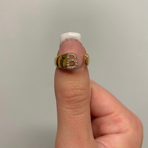 May include: A gold ring featuring a letter 'B' set with clear stones. The ring is worn on a finger.