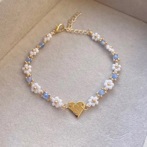 May include: A delicate bracelet featuring white daisy-shaped beads, light blue beads, and gold-coloured accents. A gold heart charm with the letter "S" is centered on the bracelet. The bracelet is on a neutral background.