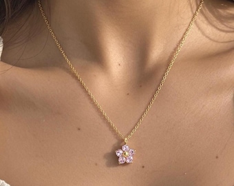 May include: A gold-toned chain necklace featuring a small flower-shaped pendant. The pendant consists of five light pink, petal-shaped stones arranged around a central gold-toned bead, creating a delicate floral design that rests against the neck.