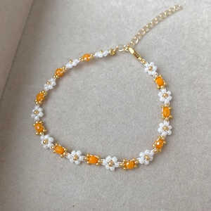 May include: A delicate gold chain bracelet with white and orange flower-shaped beads. The bracelet is made of small, evenly spaced beads.