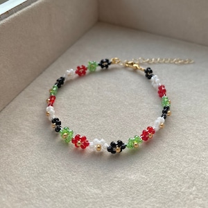 May include: A beaded bracelet with a gold clasp and a pattern of red, green, white, and black beads. The beads are arranged in a flower shape.