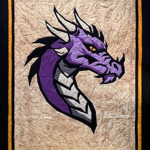 Può includere: Una trapunta rettangolare con un'applicazione centrale di una testa di drago in tonalità viola, grigio e nero con un occhio giallo. Il drago è posto su uno sfondo beige screziato, incorniciato da un bordo nero e una rifinitura gialla.