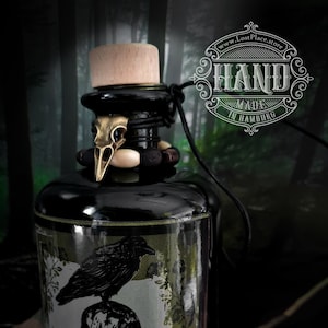 May include: A black glass bottle with a black raven and skull graphic on the label. The bottle is labeled "The Black Raven Gin & Soul" and "Lostplace Store Blackline". The bottle has a wooden stopper and a black leather cord with a raven skull charm.