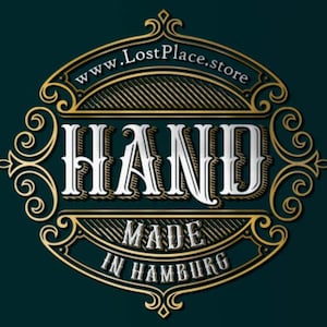 Op de afbeelding: Een gouden en wit logo met sierlijke details. Het logo zegt "HAND MADE IN HAMBURG" en "www.LostPlace.store".