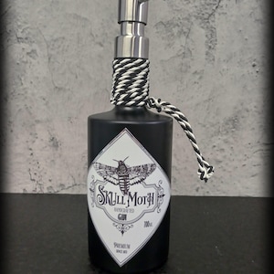 May include: Black glass bottle with a silver pump dispenser and a white and black rope tied around the neck. The bottle has a label with a black and white illustration of a moth and the text "Skull Moth Handcrafted Gin 700ml Premium Since 2013."