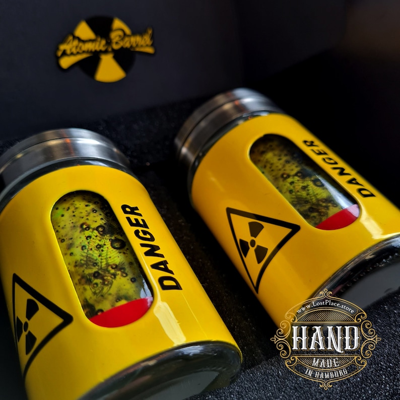 May include: Two yellow glass salt and pepper shakers with a black and white radiation symbol and the word "DANGER" printed on them. The shakers are filled with green glitter and have a red stripe around the middle. The shakers are in a black box with the words "Atomic Barrel" printed on the top.